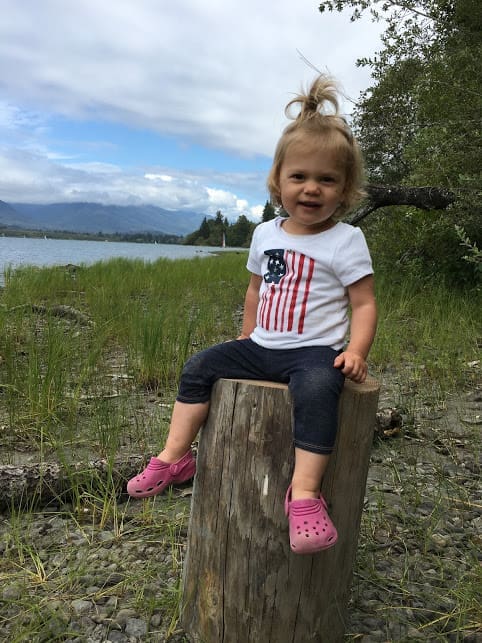 A young child wearing a white shirt with an American flag, dark pants, and pink shoes sits smiling on a tree stump by a lake, with grass, rocks, and mountains in the background.