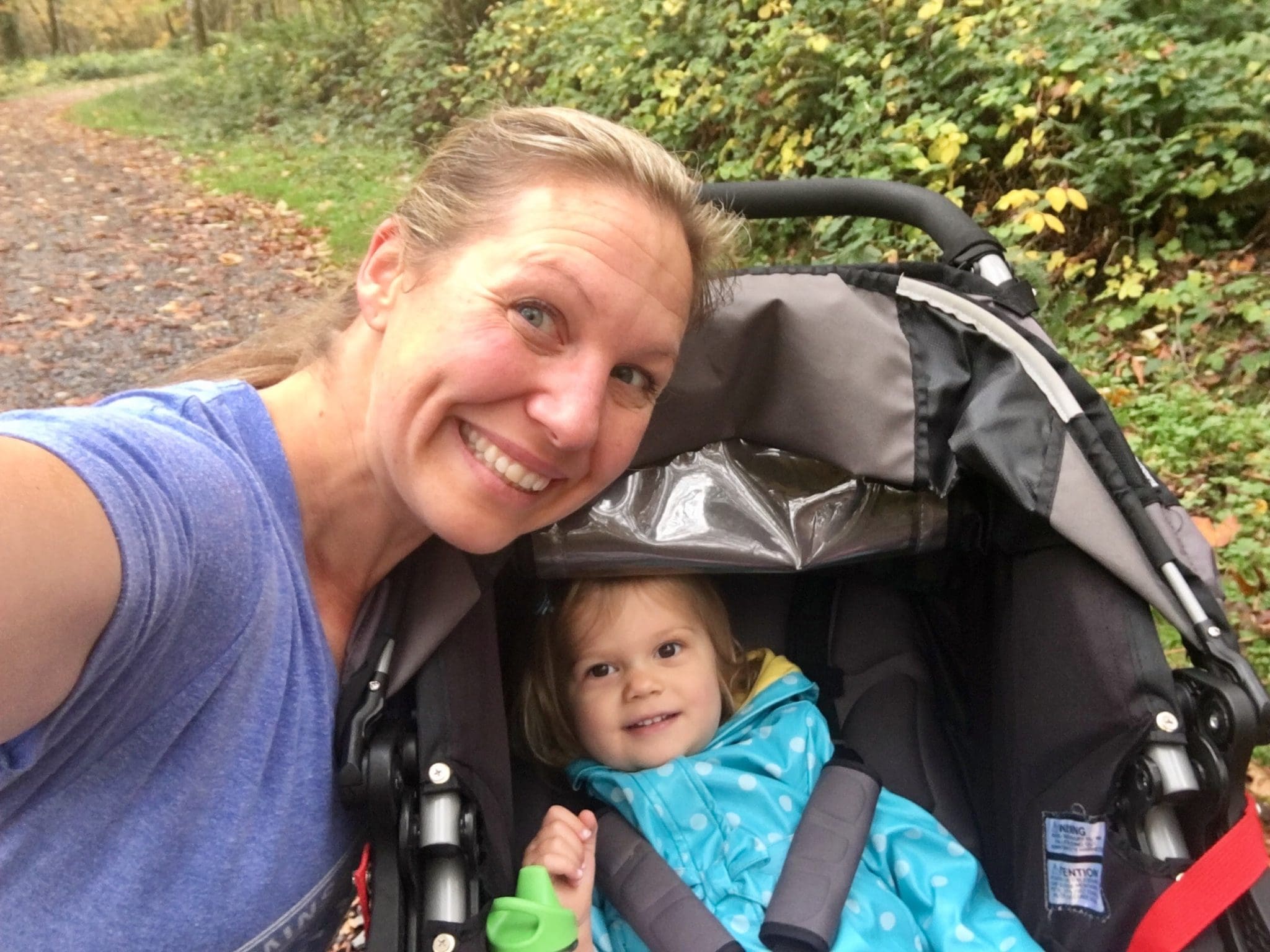 A woman smiles and takes a selfie with a young child sitting in a stroller. They are outdoors on a leafy path, surrounded by green foliage. The child wears a blue polka-dot jacket and smiles at the camera.
