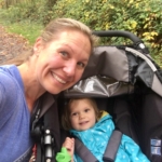 A smiling woman takes a selfie with a young child sitting in a stroller. They are outdoors on a wooded path, and the child is wearing a blue polka-dot jacket. Greenery surrounds the path in the background.