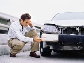 A man crouches next to a damaged car, examining the broken front bumper with a concerned expression while talking on his phone, as the distant roar of Blue Angels jets echoes from a nearby Seafair event. The car is missing part of its front and headlight.