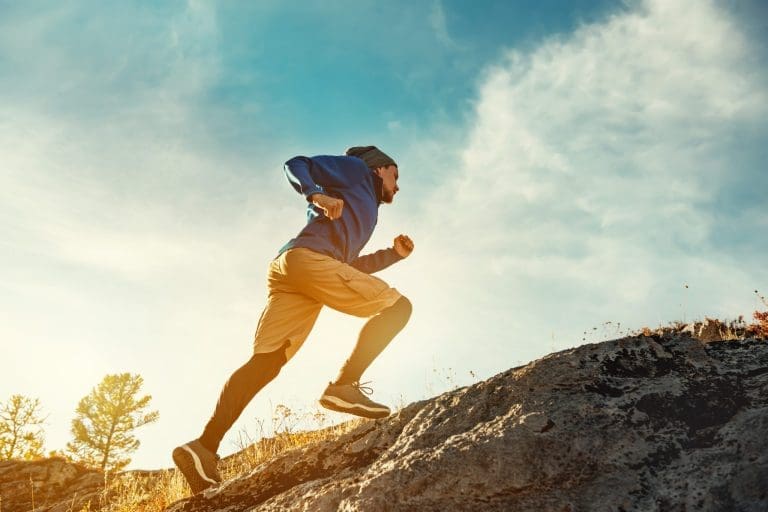 A person demonstrating strong General Adaptive Potential (GAP) wears athletic clothes and a beanie as they run uphill on rocky terrain outdoors, with a bright blue sky and sunlight in the background.