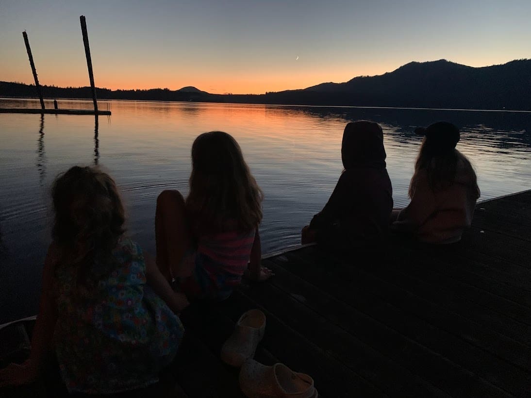 Four children sit on a dock at dusk, watching the sunset over a calm lake with mountains in the background—one of their favorite things. Their silhouettes are reflected peacefully on the water.