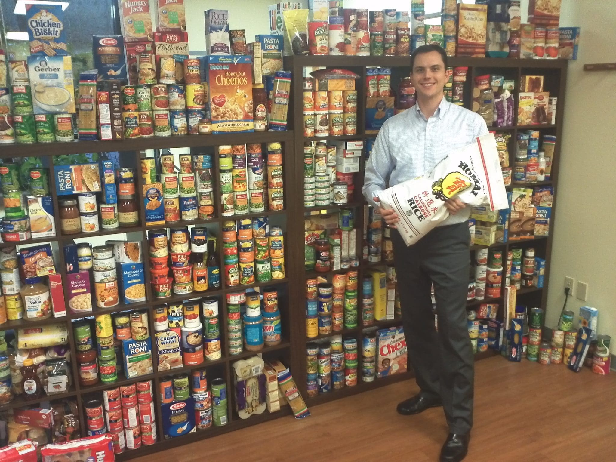 A man in business attire stands smiling beside shelves filled with canned goods and boxed foods. Holding two bags of rice, he supports the Helping Hands Food Drive in a room with a wooden floor.