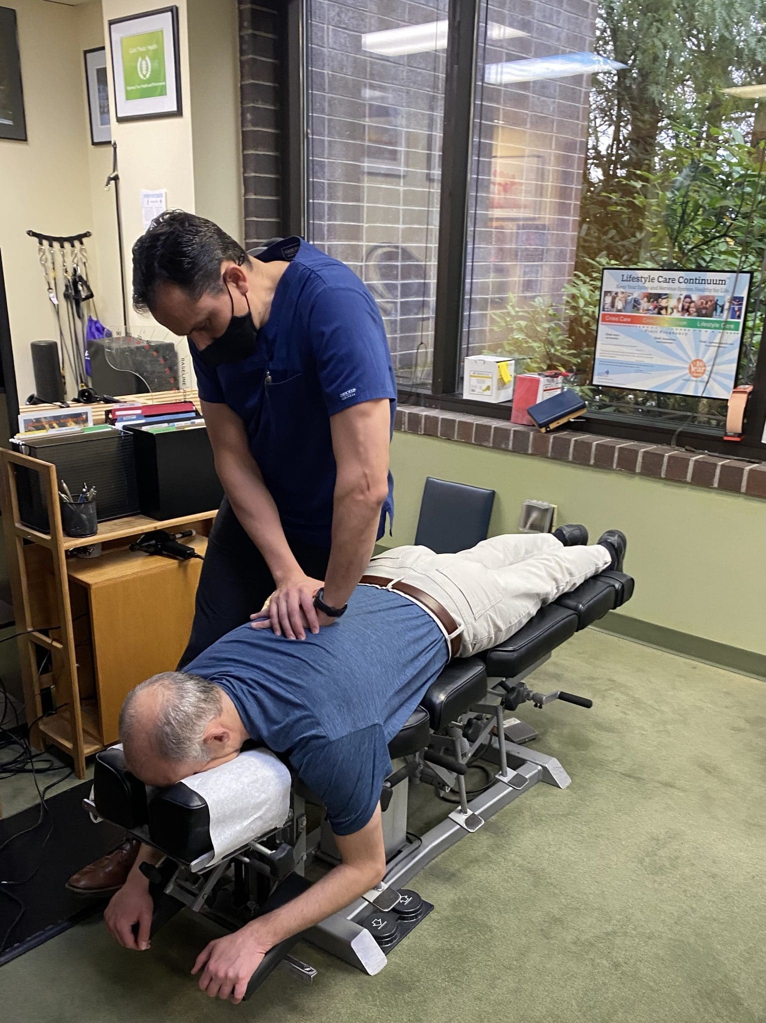 A chiropractor in blue scrubs and a face mask adjusts the back of a man lying face down on a treatment table, giving you an idea of what to expect during an office visit with large windows and various decorations.