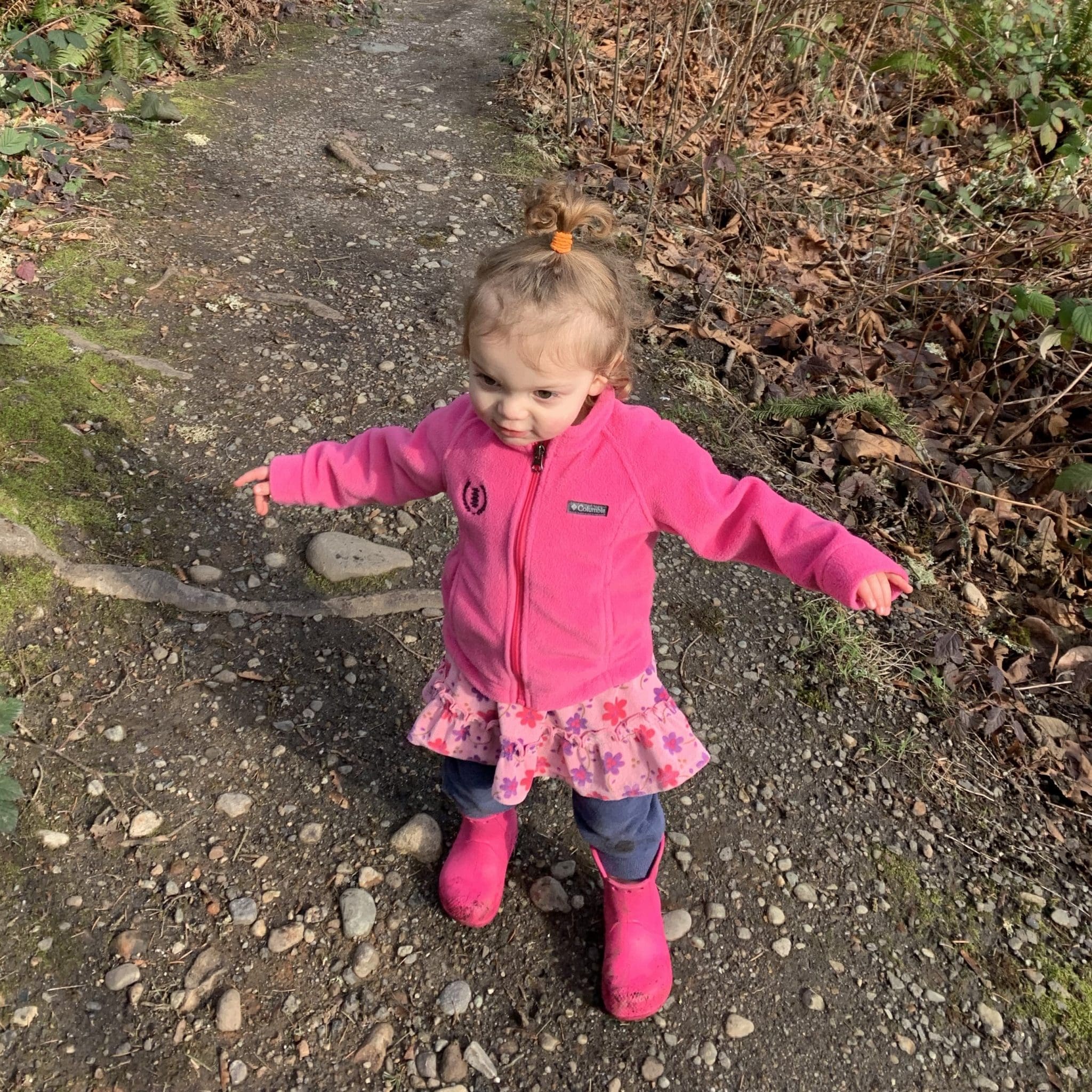 A small child with light brown hair in a top bun stands pain free on a rocky path outdoors, wearing a bright pink jacket, pink floral skirt, and pink rain boots, with arms outstretched. Dry leaves and moss surround the path.