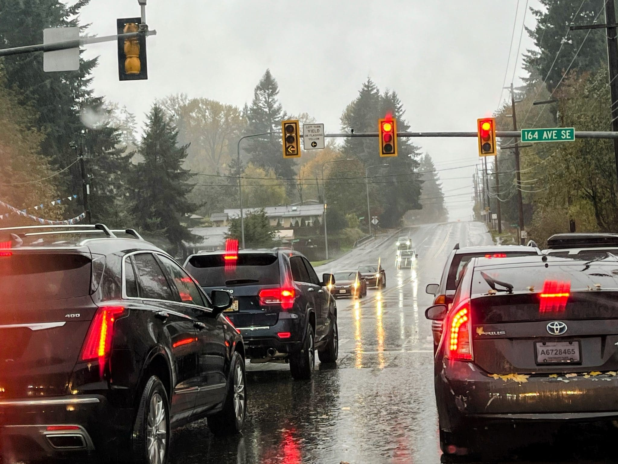 Cars wait at a red traffic light on a rainy day at the intersection of 164 Ave SE. Reflections from taillights shimmer on wet pavement, while trees line both sides—a quiet moment to recover after an earlier accident on the road.