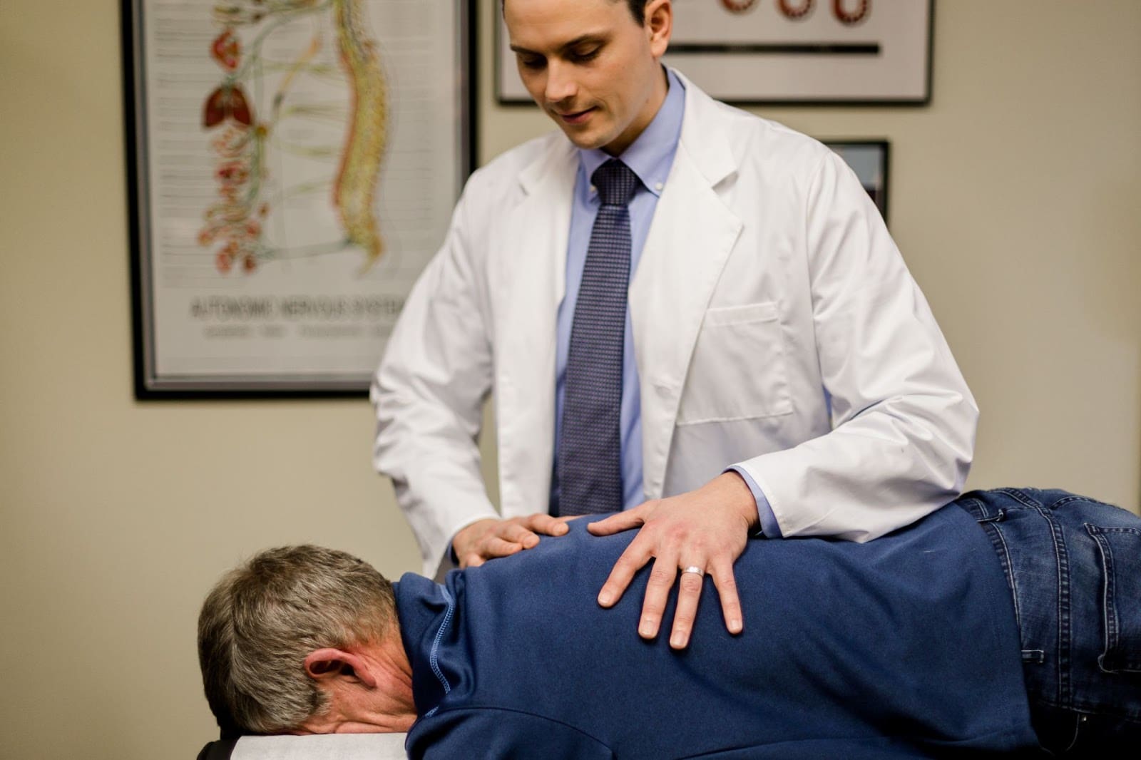 A doctor in a white coat examines a patient lying face down on an exam table, placing hands on the patient's back—an important step in assessing recovery from whiplash. Medical charts are visible on the wall in the background.