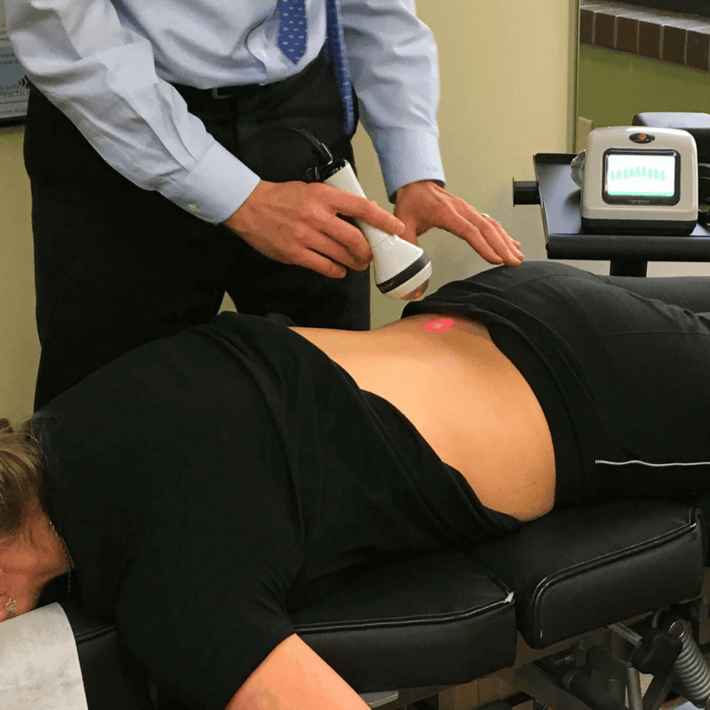 A person lies face down on a therapy table while a practitioner uses a handheld device to provide laser therapy for pain on their lower back. A small red light is visible on the skin, and medical equipment is nearby.