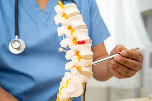 A healthcare professional in blue scrubs holds a pen and points to a model of a human spine, possibly explaining spinal conditions or treatment options related to posture correction.