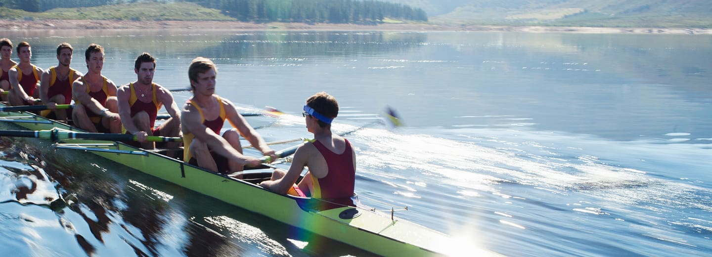 About eight rowers in matching uniforms power their long boat in perfect unison across a calm lake, surrounded by trees and hills on a bright, sunny day.