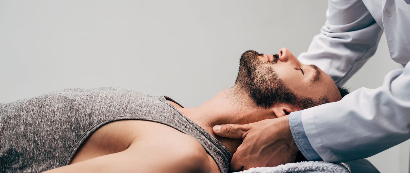 A man with a beard lies on his back while a Car Accident Chiropractor in a white coat gently examines his neck, suggesting a therapeutic treatment session.