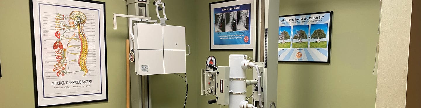 A patient center chiropractic office with an X-ray machine, anatomical spine chart, and spinal health posters on green walls. The clean, organized space keeps medical equipment visible for patient comfort and care.