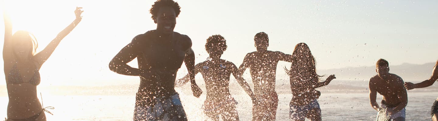 A group of people in swimwear laughing and playing in contact with the water at the beach, sunlight shining behind them and water splashing around.