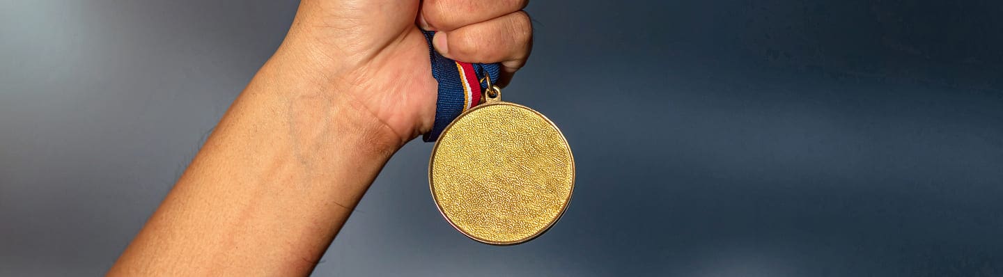 A hand holds up a gold medal with a blue, red, and white ribbon against a dark, blurred background—symbolizing achievement after recovery with the help of a Car Accident Chiropractor.