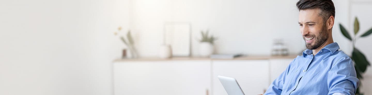 A smiling man in a blue shirt sits indoors using his laptop to browse the FAQ section. The background is softly lit with minimal decor, featuring plants and shelves.