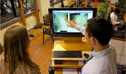 A man and a woman stand by a computer monitor in a patient center as the man points to medical images on the screen in a well-lit office with wooden floors and large windows.