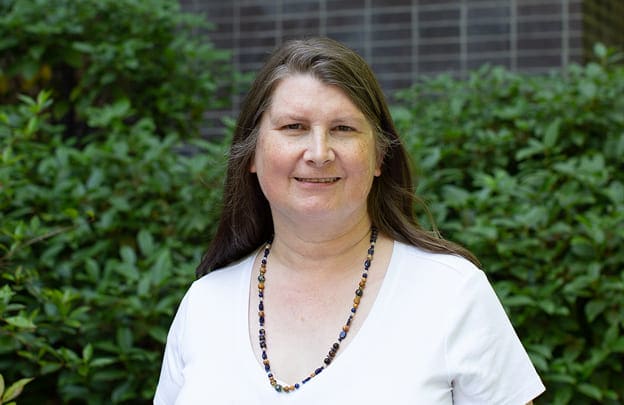 A woman with long brown hair wearing a white shirt and beaded necklace stands outdoors in front of green bushes and a brick wall, smiling at the camera.