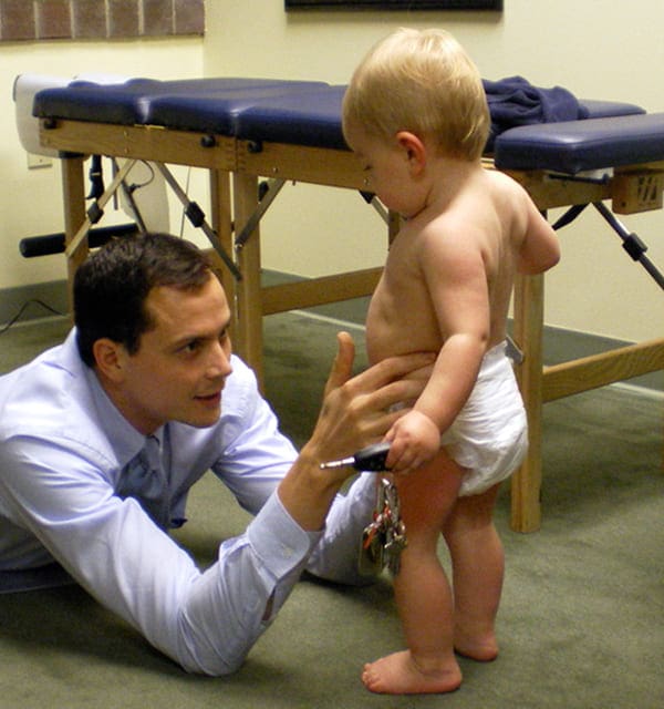 A man crouches on the floor, smiling and reaching out to a toddler in a diaper who is standing nearby, while the man—possibly a car accident chiropractor—holds a set of keys. A medical exam table is visible in the background.