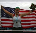 A female athlete wearing a medal holds a bouquet and spreads an American flag behind her, smiling in celebration.