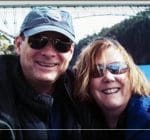 A man and woman wearing sunglasses smile together outdoors, with a bridge and trees visible in the background. The man wears a cap and jacket, and the woman has shoulder-length hair and a blue scarf.