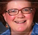 A person with short, curly brown hair and glasses smiles at the camera. They are wearing patterned earrings and a blue top, with a blurred dark background.