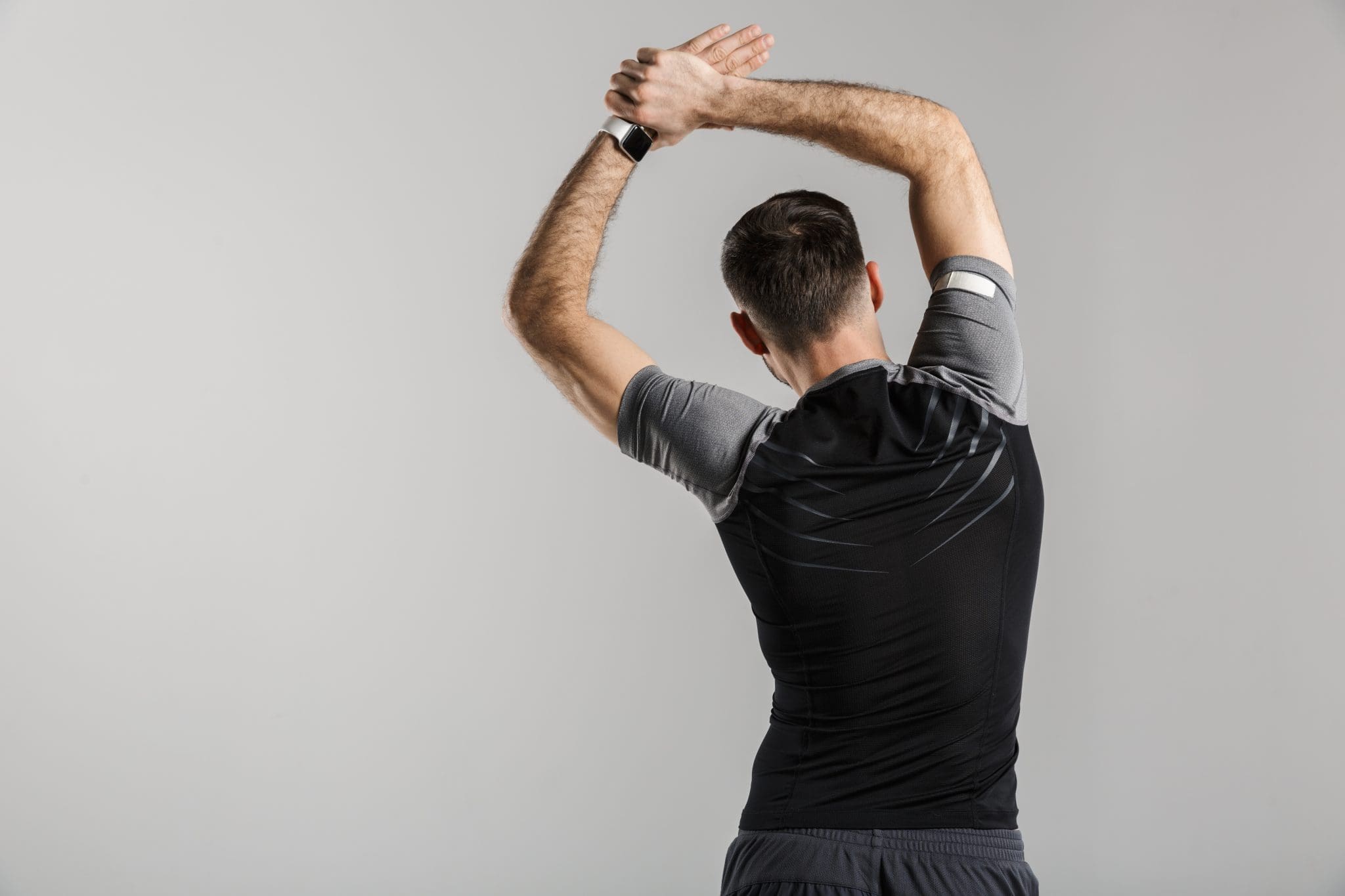 A man in athletic clothing stands with his back to the camera, stretching his arms overhead and to the side against a plain, light gray background, demonstrating a simple move for better spinal hygiene.