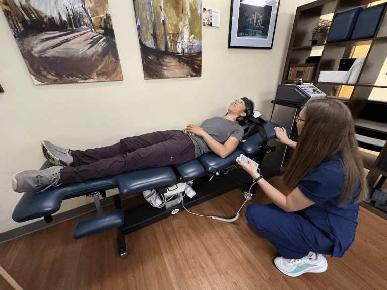 A person lies on a chiropractic adjustment table while a healthcare professional in scrubs kneels beside them, possibly demonstrating spinal decompression vs traction. The room features wooden floors, paintings, and various medical equipment.