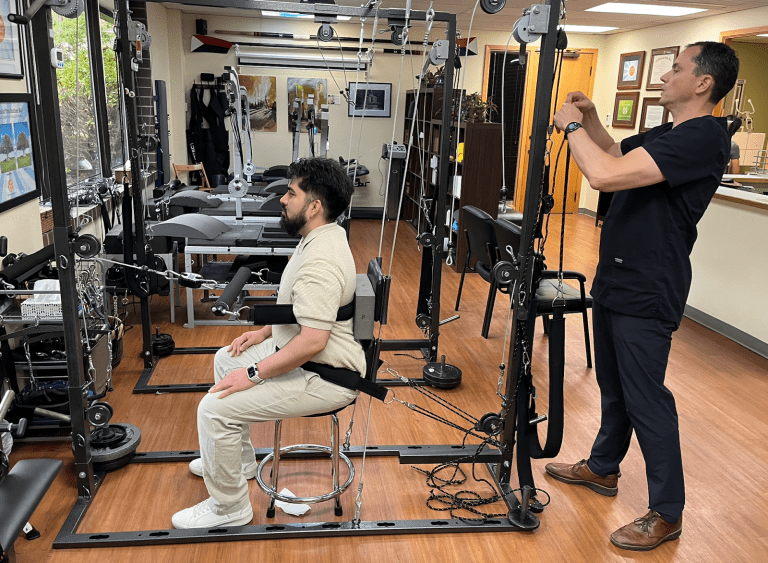 A man sits on a stool inside a physical therapy machine, possibly used for spinal decompression vs traction, while a therapist adjusts cables. The room features exercise equipment, framed certificates, and wood flooring.