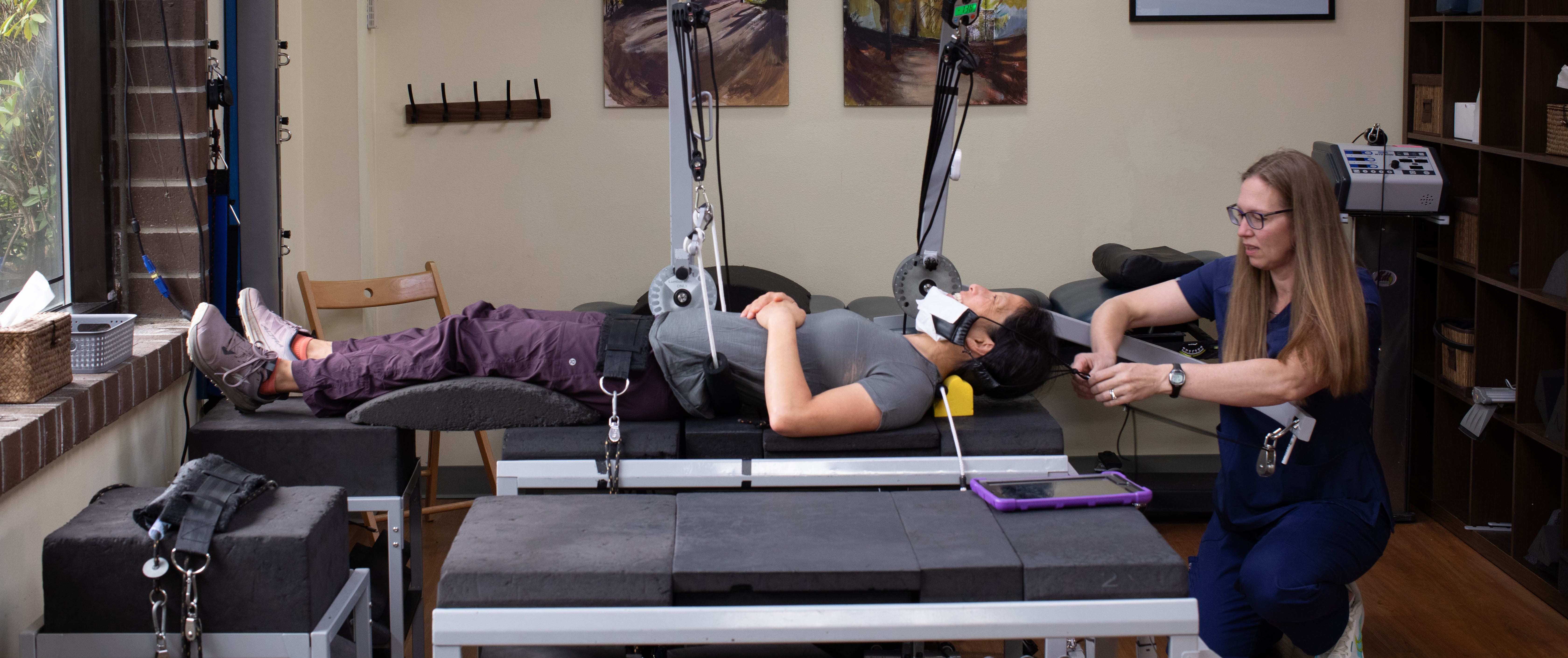 A woman lies on a therapy table with devices attached to her head and chin, while a Car Accident Chiropractor adjusts the equipment in what appears to be a medical or physical therapy clinic.