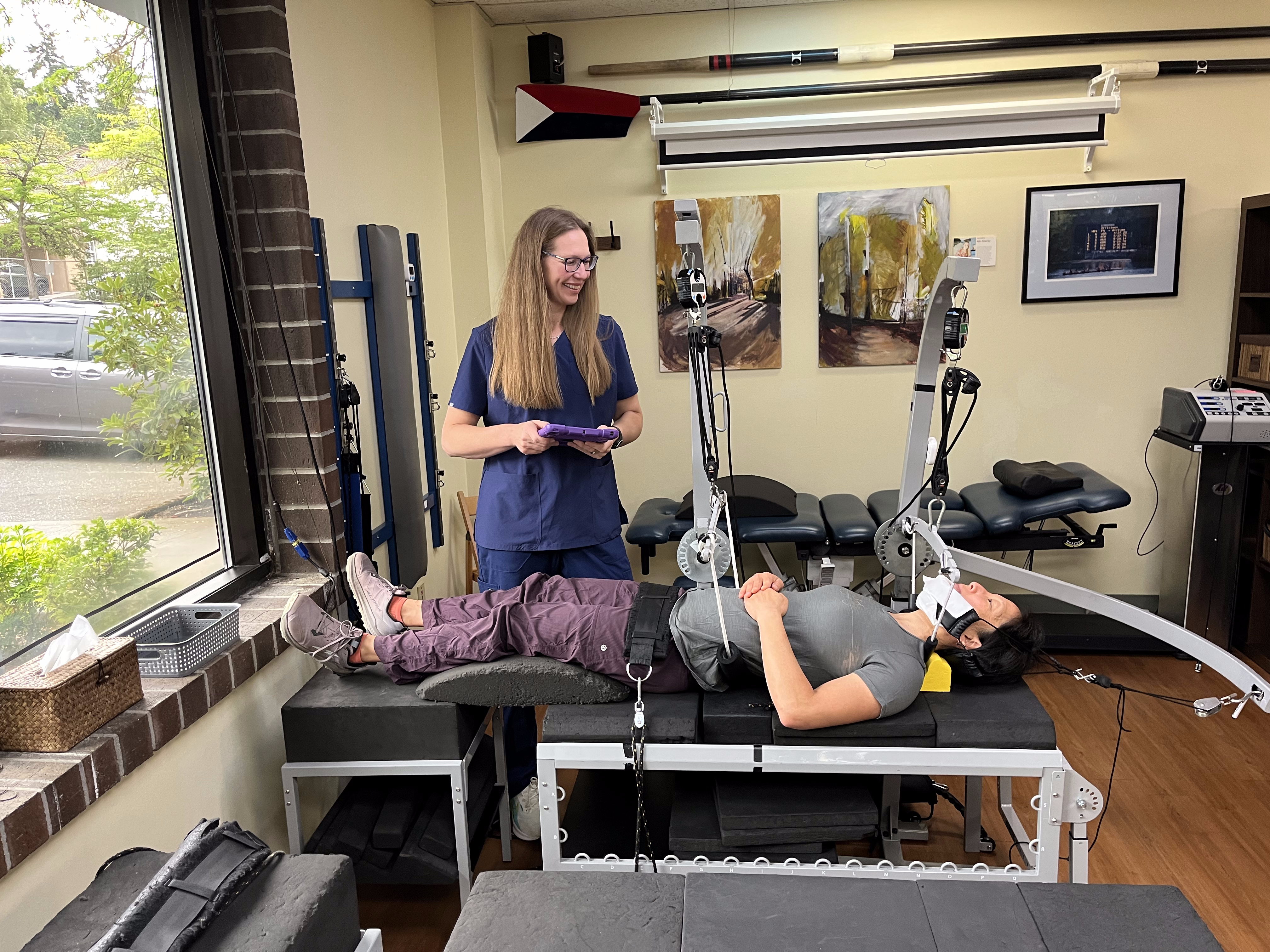 A physical therapist stands beside a patient lying on a black treatment table, connected to a headache treatment device with straps on her legs. The room features medical equipment, artwork, and a large window offering an outdoor view.