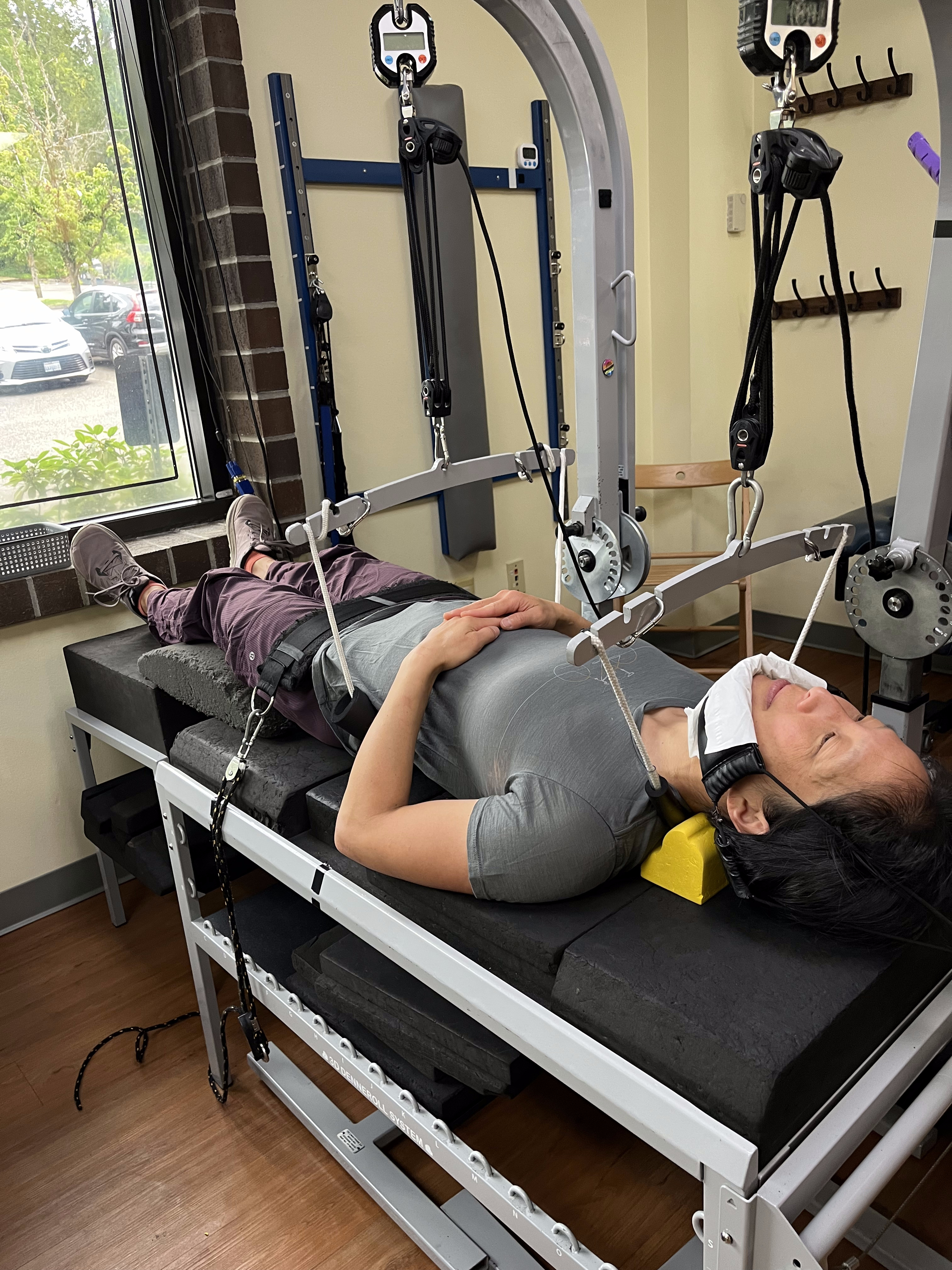 A person is lying on a therapeutic table indoors, using a suspension system with straps and pulleys for neck and back support as part of a headache treatment. They wear a neck brace and rest with hands on their abdomen near a window.