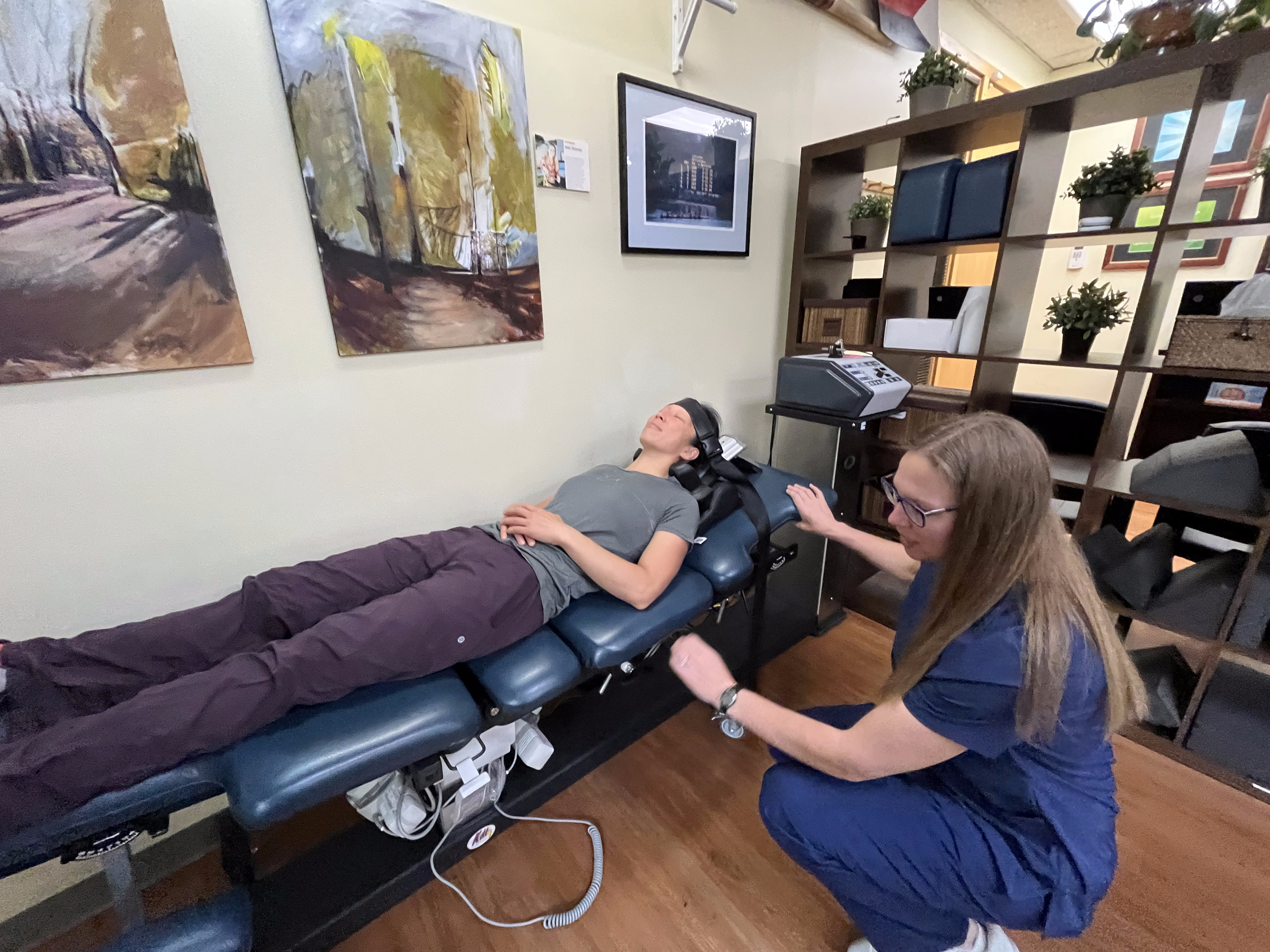 A person lies on a chiropractic adjustment table for headache treatment as a practitioner in scrubs prepares to treat them. The room features paintings on the wall, a shelf with plants, and nearby medical equipment.