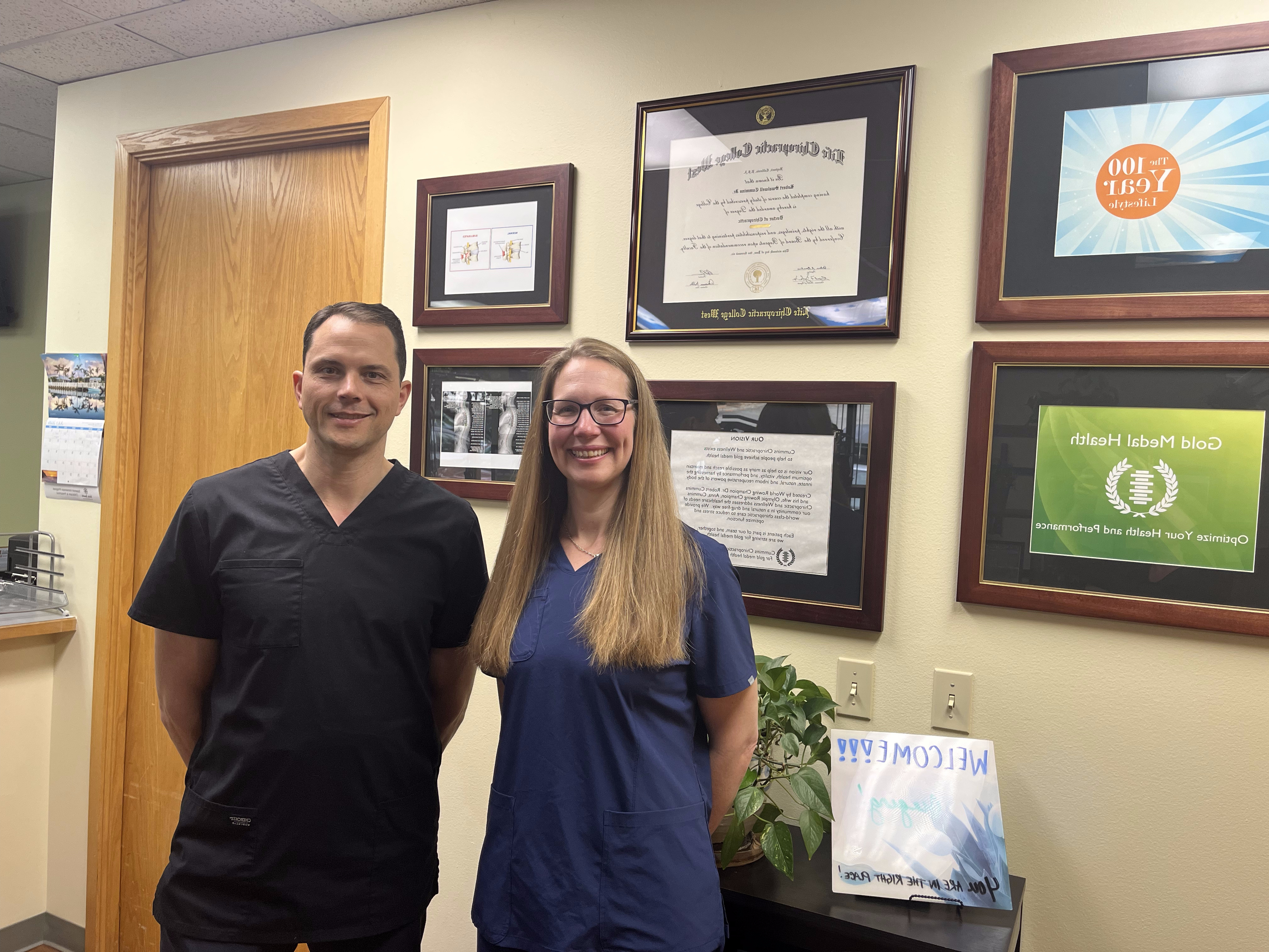 Two healthcare professionals in scrubs stand smiling in an office decorated with framed certificates and health-related posters, including information about headache treatment. A Welcome sign and plant add a friendly touch to the nearby table.