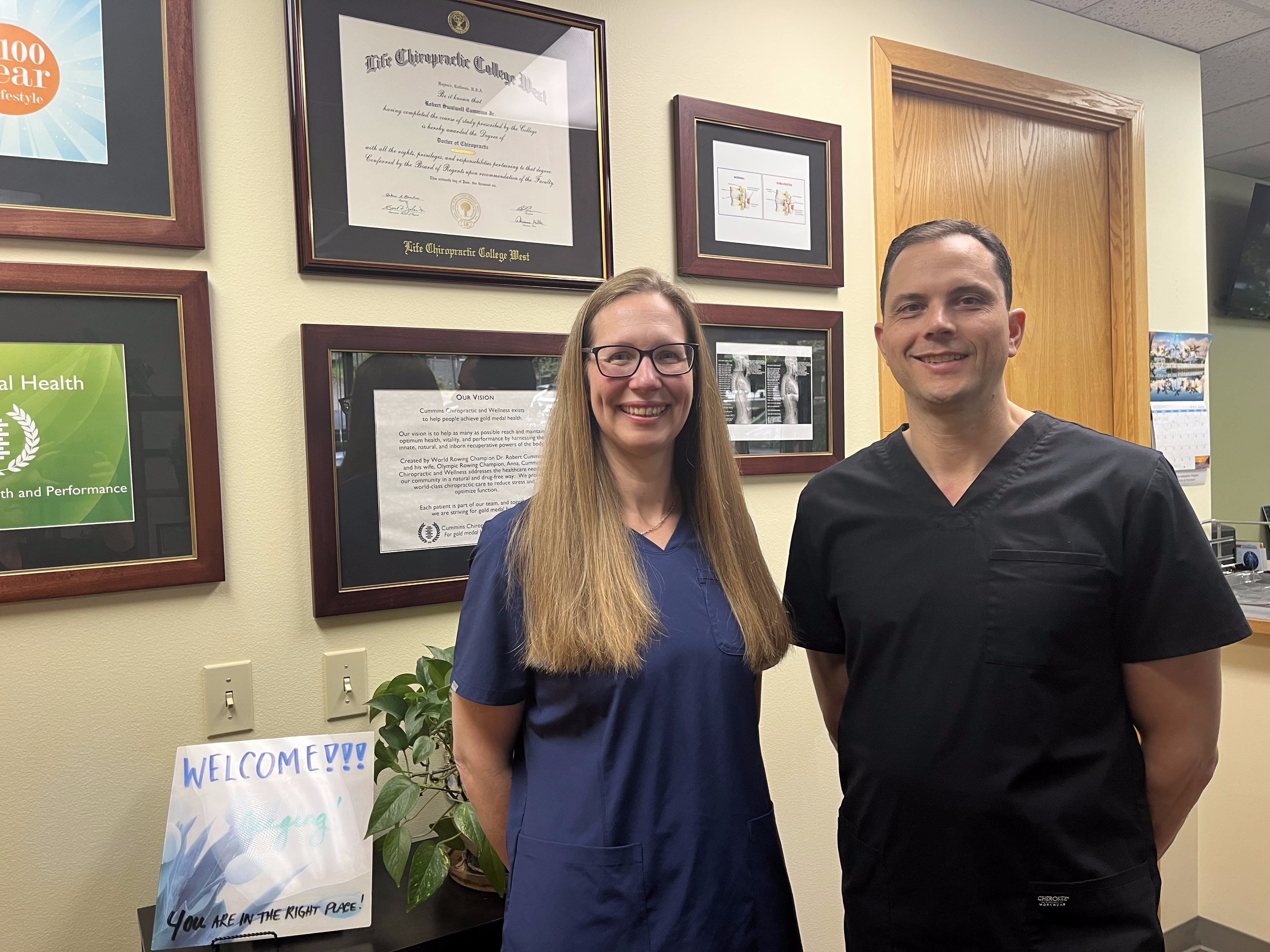 Two people in dark scrubs stand smiling in an office with framed certificates and awards behind them. A “Welcome” sign and a potted plant are visible, creating a warm space specializing in Headache Treatment.