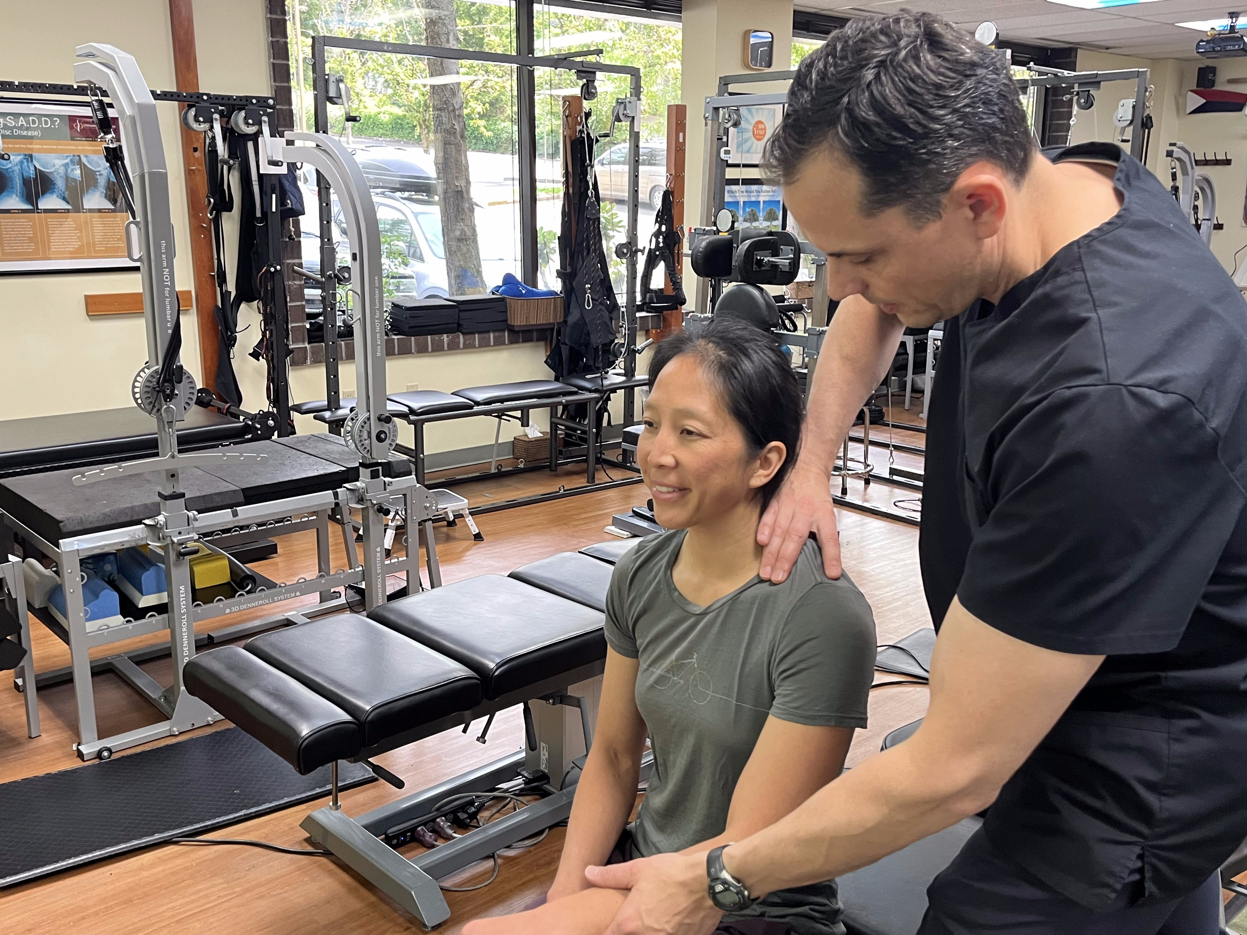 A physical therapist in black scrubs evaluates a woman’s shoulder in a gym-like clinic, which also offers headache treatment, with exercise equipment and large windows in the background. The woman sits on a bench, looking forward and smiling slightly.