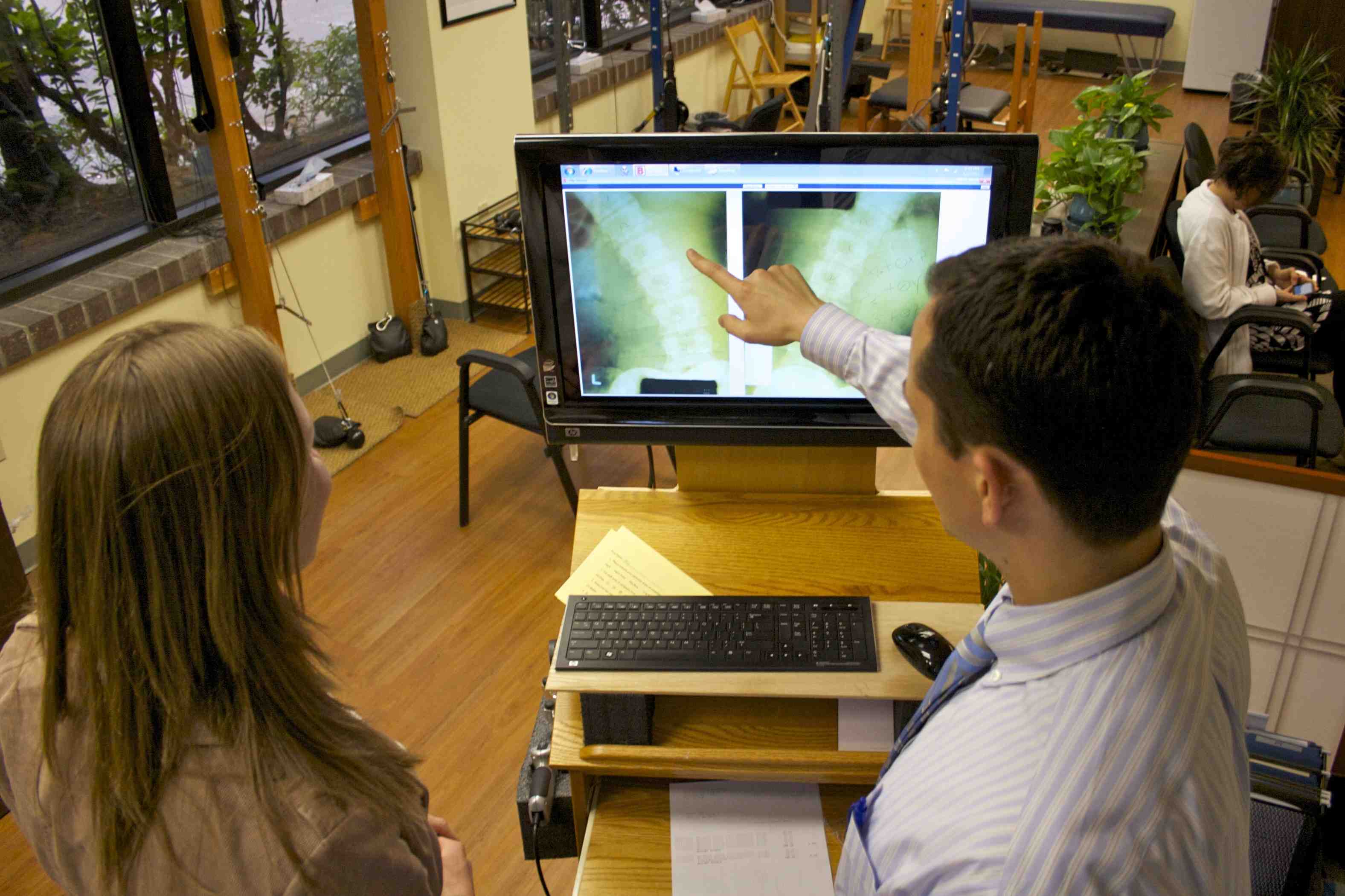 A doctor discusses X-ray images related to headache treatment with a patient in a medical office featuring wooden floors and large windows.
