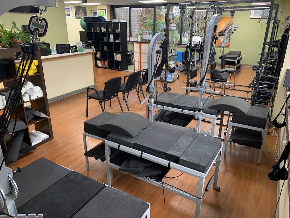 A physical therapy clinic offering forward head posture treatment, with exercise tables, black cushioned equipment, chairs, and shelves with plants and books. Large windows fill the room with light, highlighting the wood floors and green walls.