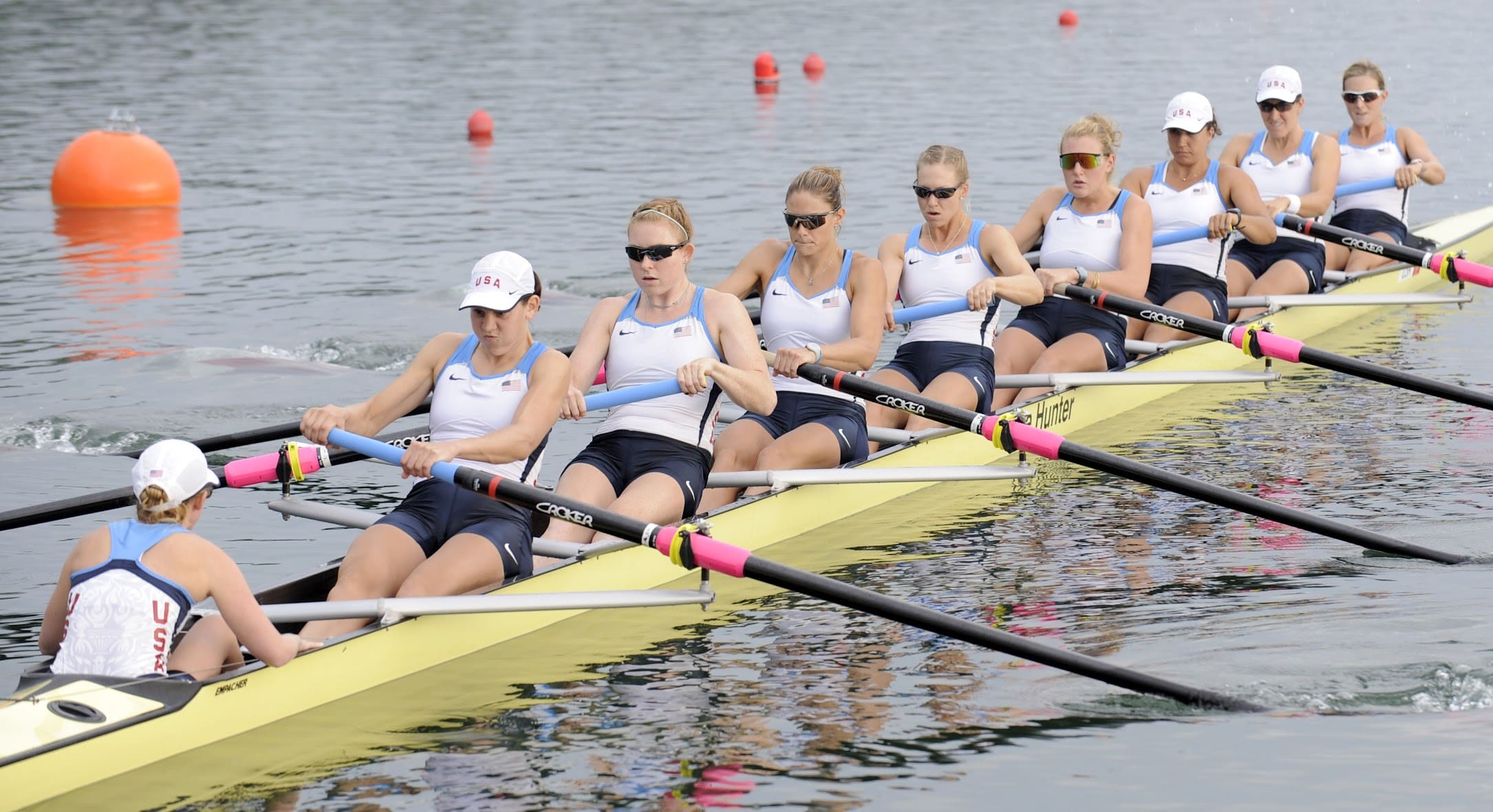 Eight female rowers in matching uniforms and sunglasses, along with a coxswain, power their yellow racing shell down calm water during a competition, where peak performance and sports chiropractic care can make all the difference. Red buoys mark their lane.