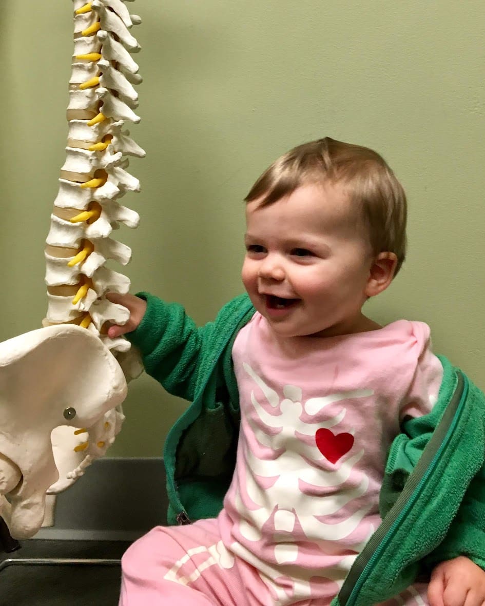 A smiling baby in a skeleton-themed outfit sits beside a model spine and pelvis, with a green jacket draped over their shoulders, evoking the playful spirit of a pediatric chiropractor, all set against a pale green wall.
