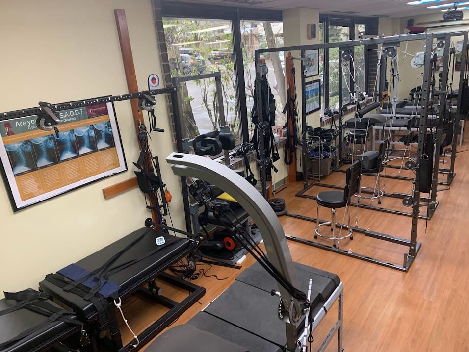 A rehabilitation or physical therapy clinic offering Postural Distortion Treatment, with various exercise and therapy equipment, including tables, resistance bands, pulleys, chairs, and large mirrors. The room has wooden floors and ample natural light from large windows.