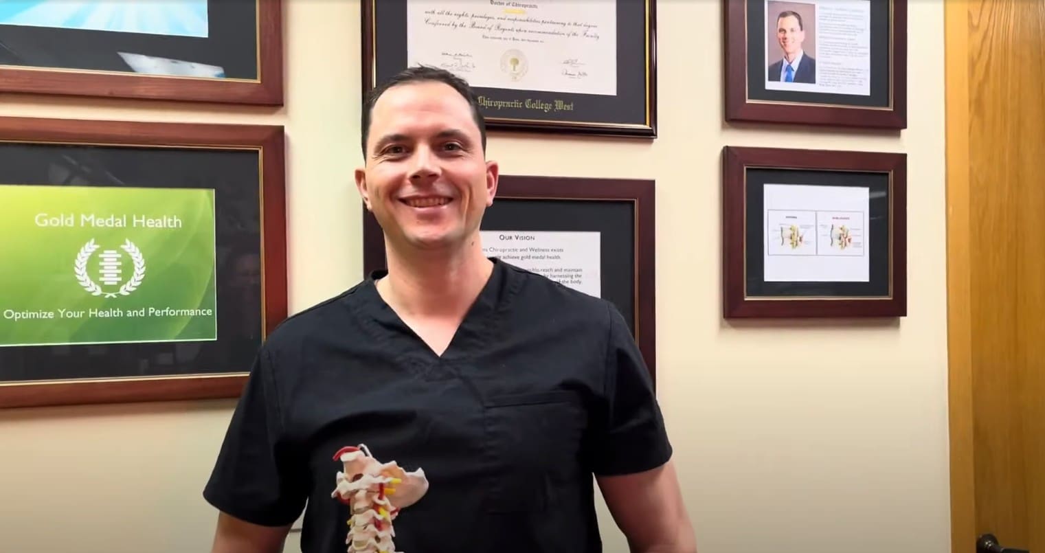 A man in black scrubs stands in an office, smiling and holding a model of a spine—reflecting his passion for chiropractic care. Framed certificates, documents, and a photo are displayed on the wall behind him.