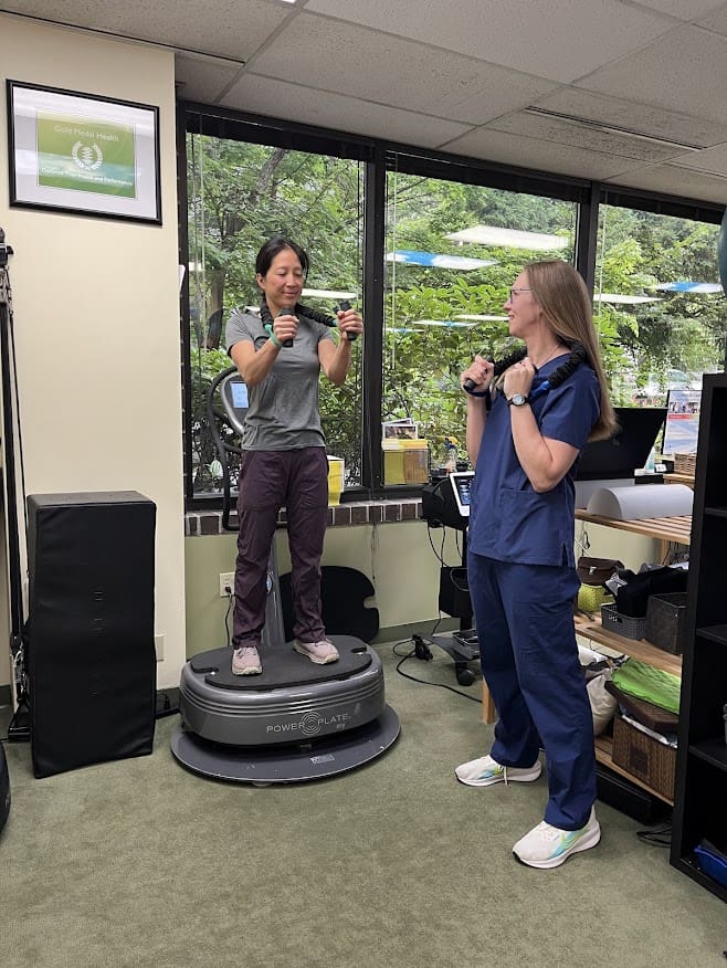 Two women exercise in an office. One stands on a vibration plate holding dumbbells, while the other, wearing scrubs, stands beside her also holding dumbbells. They smile in a bright room with fitness equipment for forward head posture treatment.