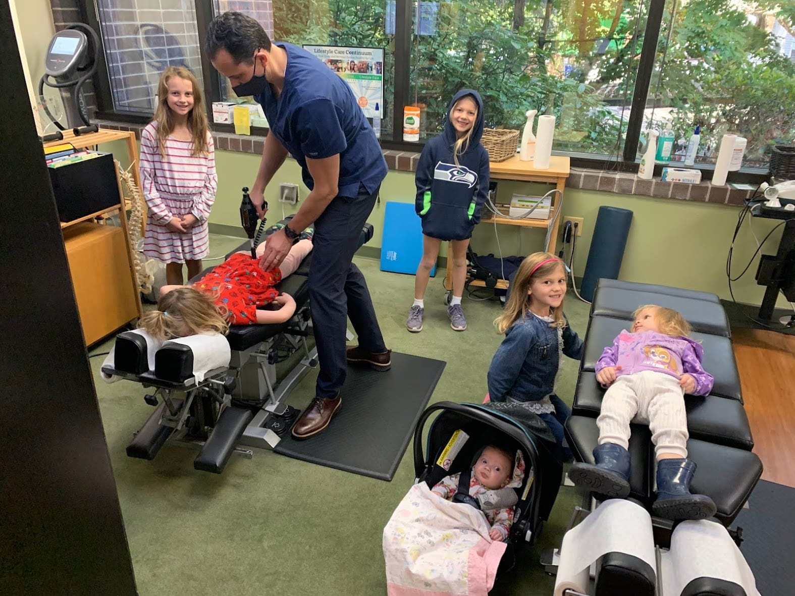 A pediatric chiropractor adjusts a young girl lying face down on a table while four other children, including a baby in a car seat, wait nearby in a brightly lit office with large windows and greenery outside.