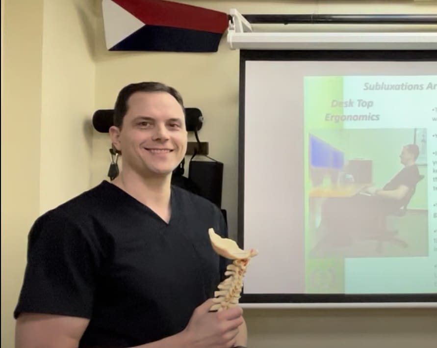 A man in medical scrubs holds a model of a human spine and jaw while standing in front of a projector screen displaying information about desk ergonomics and headache treatment. A flag is visible on the wall above.