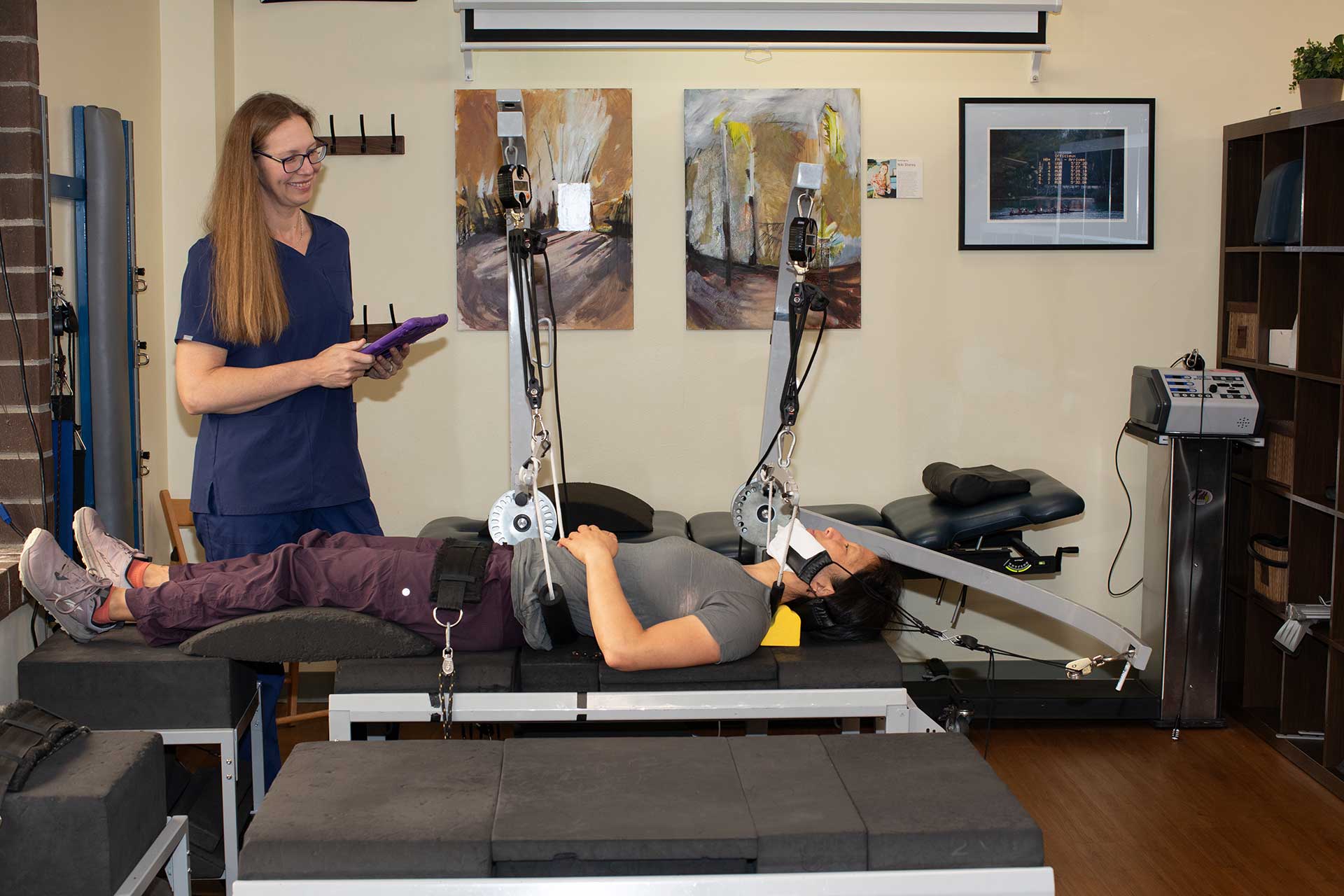 A woman lies on a table with a neck traction device attached to her head, while a chiropractor Bellevue professional in scrubs stands nearby holding a tablet and smiling. The room contains therapy equipment and artwork on the walls.