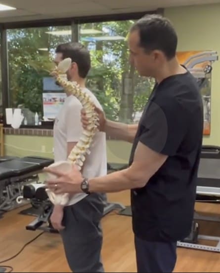 A doctor in black scrubs holds a plastic model of a human spine next to a standing patient’s back, demonstrating Hyperkyphosis Treatment in a modern medical office with exam tables and large windows in the background.