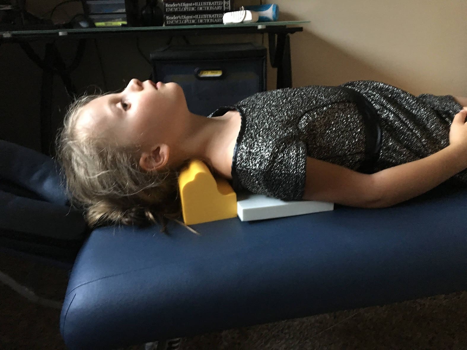 A young girl in a sparkly dress lies on her back on a blue padded table, her head resting on a yellow foam neck support during a pediatric chiropractor visit. She gazes upward, and a desk with various items is visible in the background.