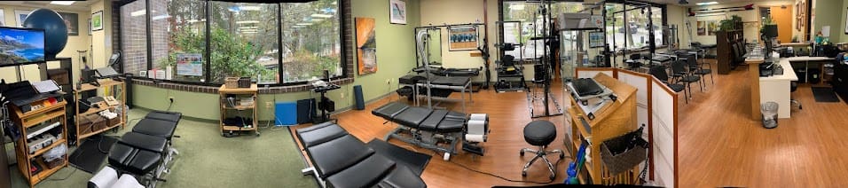 A panoramic view of a chiropractic clinic with multiple black treatment tables for Postural Distortion Treatment, exercise equipment, wooden floors, large windows, green and beige walls, and various medical devices and supplies neatly organized.