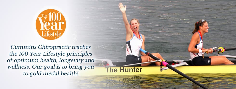 Two female rowers in a yellow boat labeled The Hunter celebrate on the water; one raises her arm in victory. Text promotes Cummins Chiropractic, sports chiropractic care, and the 100 Year Lifestyle for health and longevity.