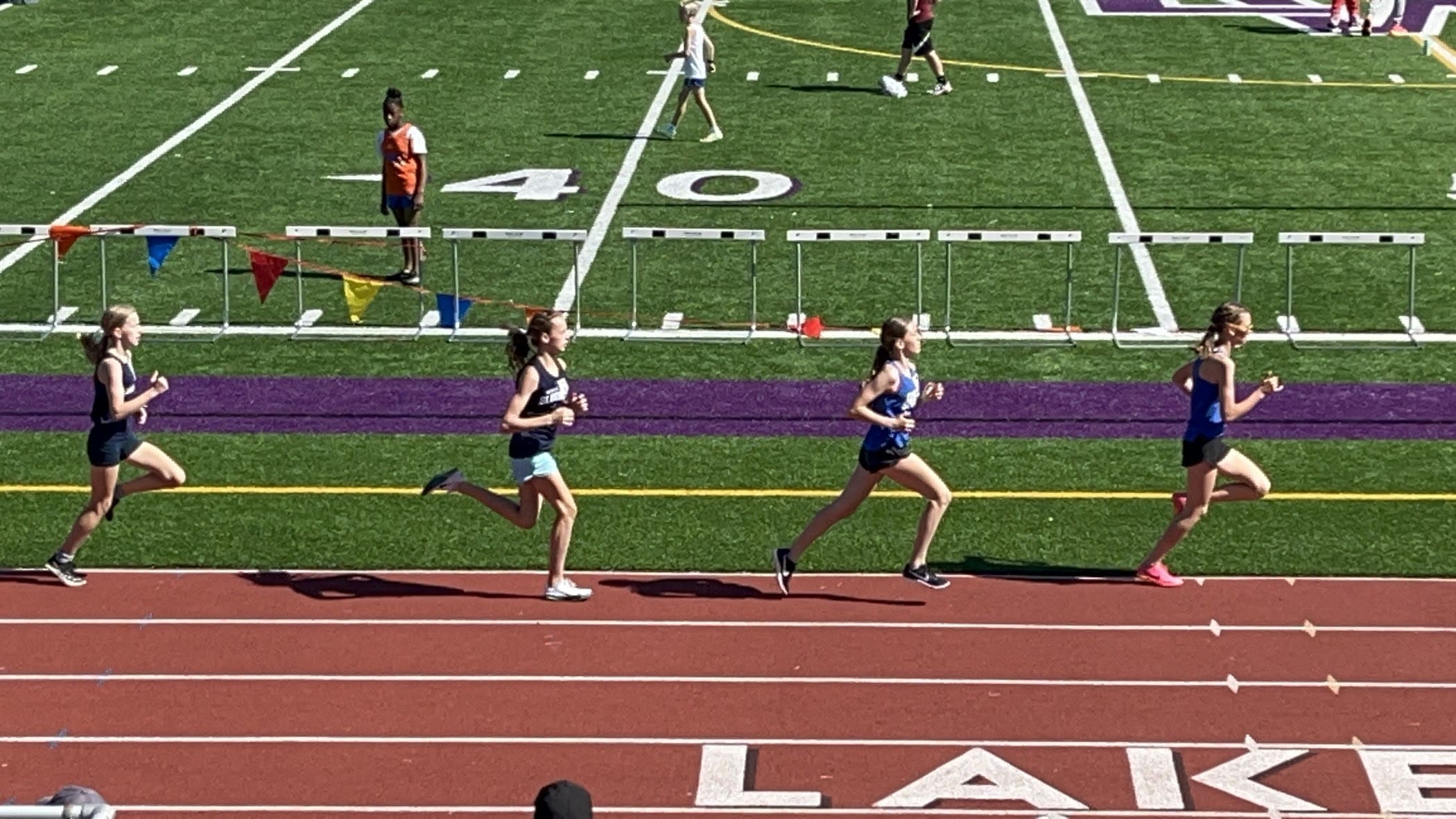Four female track runners race on a red track beside a grassy field marked with a 40-yard line. Three are closely grouped, with one slightly ahead, demonstrating the peak performance that sports chiropractic care can help support. Spectators look on in the background.